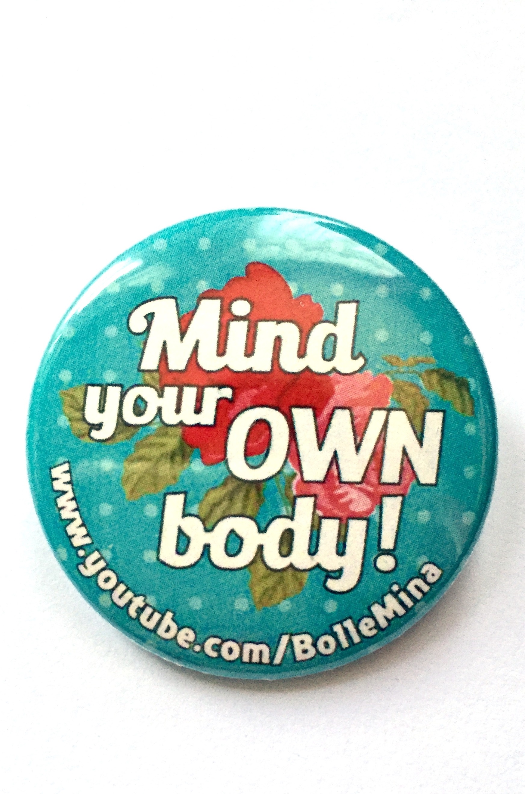 Foto van een button mt een turquoise achtergrond met lichtblauwe stipjes. Hierop een grote rood roze roos en in witte letters de tekst Mind your own body 